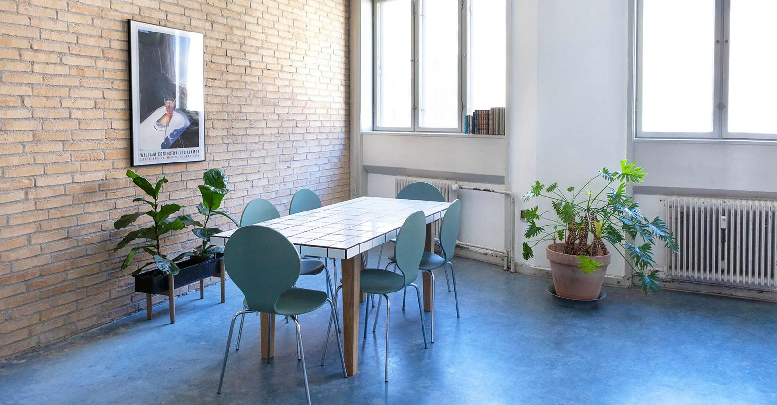 A long table with green chairs in front of a yellow brick wall and tall windows.